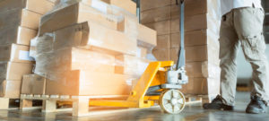 Young man with hand pallet truck or pallet jack and stack of cardboard boxes on pallet in distribution warehouse.