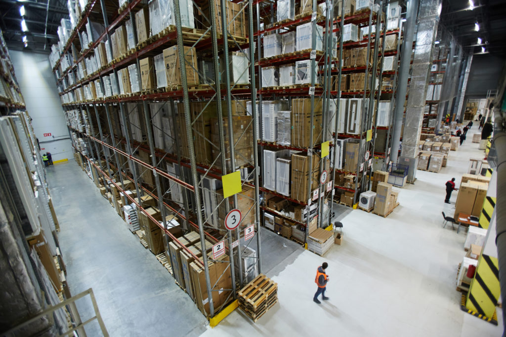 Large factory or warehouse with workers monitoring goods stacked on shelves from floor to ceiling.