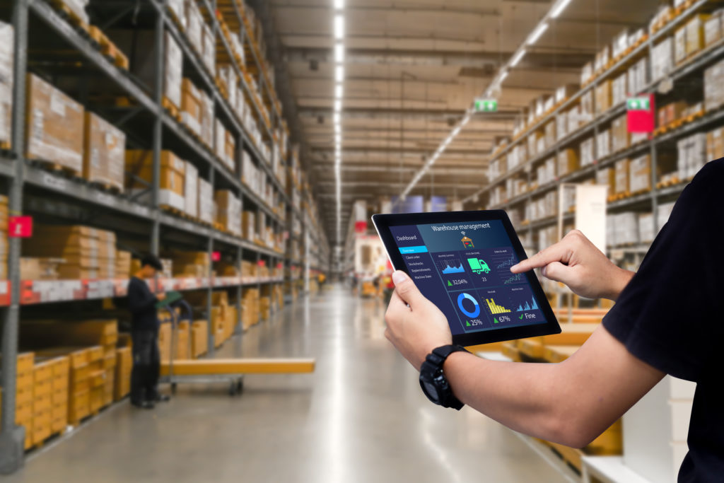 An employee consults a tablet while looking out into a full warehouse stocked with inventory.